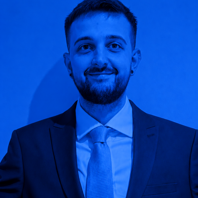 Luke26 Professional headshot of a man in a dark suit and light blue tie, smiling at the camera.