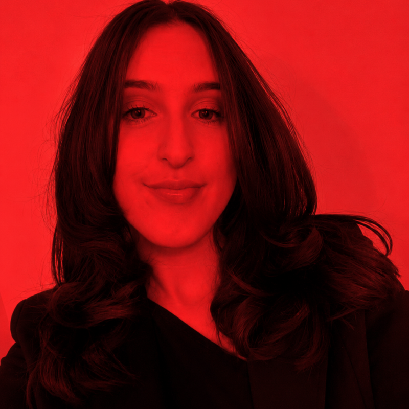 Mel26 Portrait of a woman with shoulder-length dark hair wearing a black blazer, smiling softly against a red background.